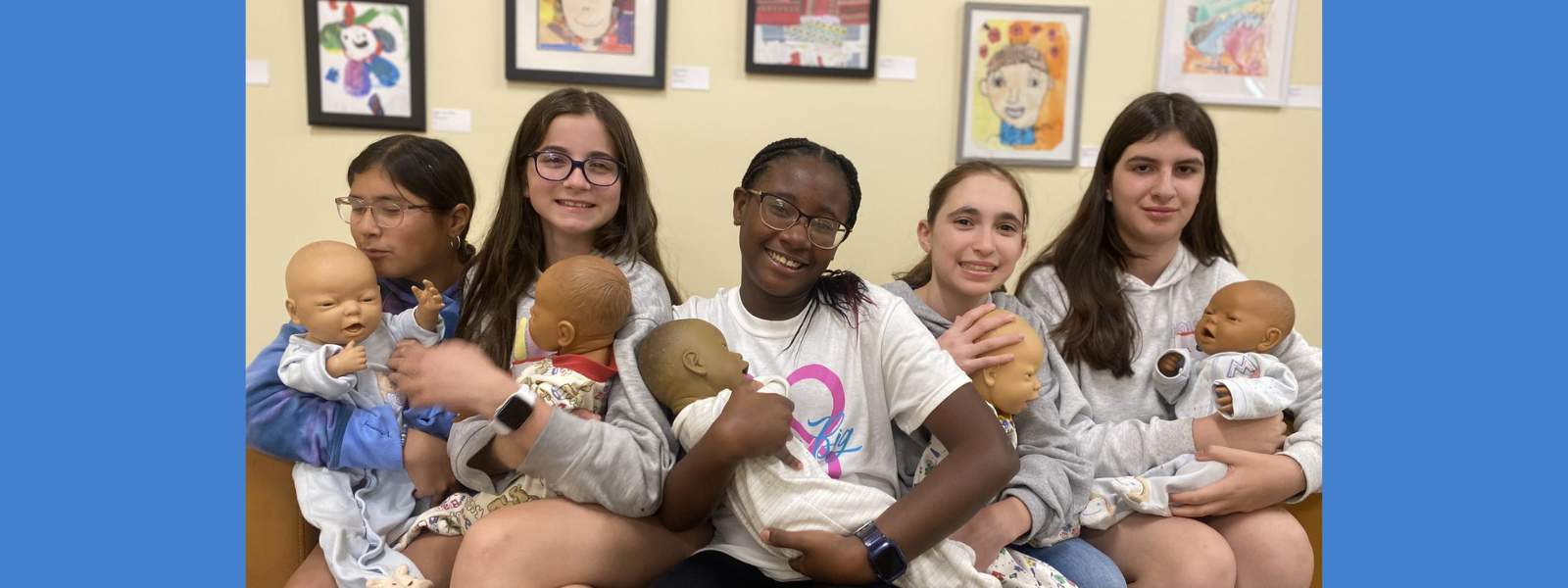 Five girls smile while holding baby dolls in front of colorful framed artwork. The mood is cheerful and playful.