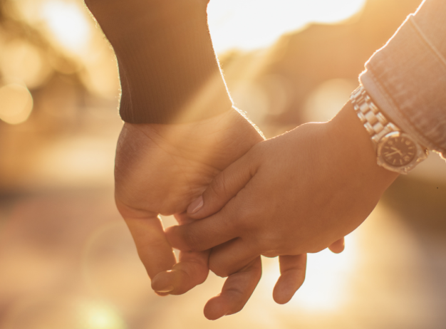 Two people holding hands outdoors, with sunlight shining in the background. One person wears a watch and a denim jacket; the other has on a dark long-sleeve shirt.
