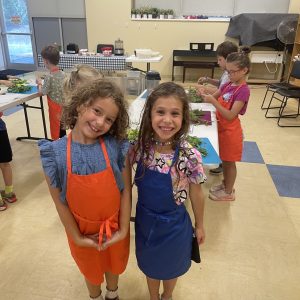 Two young girls smile at the camera while wearing aprons in a classroom or kitchen setting. Other children in the background are preparing food at tables. The room has light walls and floors, with windows and a clock on the wall.