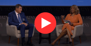 Two people seated on stage in a talk setting, engaged in discussion. The person on the left in a blue suit, and the person on the right in an orange dress.