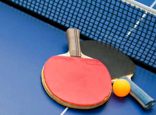 Two table tennis paddles, one red and one black, and an orange ping pong ball rest on a blue table next to the net.