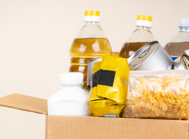 A cardboard box filled with food supplies, including pasta, canned goods, cooking oil, and water bottles, is being held by someone wearing a gray sweater.