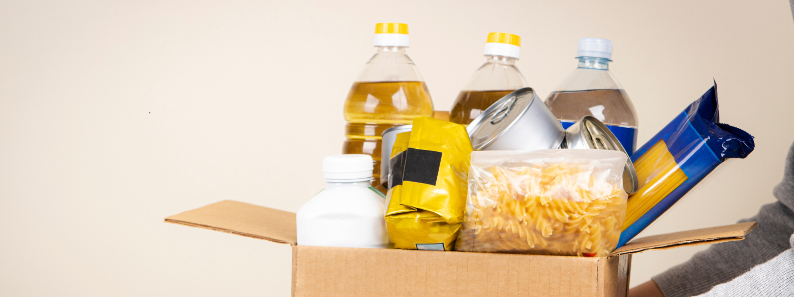 A cardboard box filled with food supplies, including pasta, canned goods, cooking oil, and water bottles, is being held by someone wearing a gray sweater.