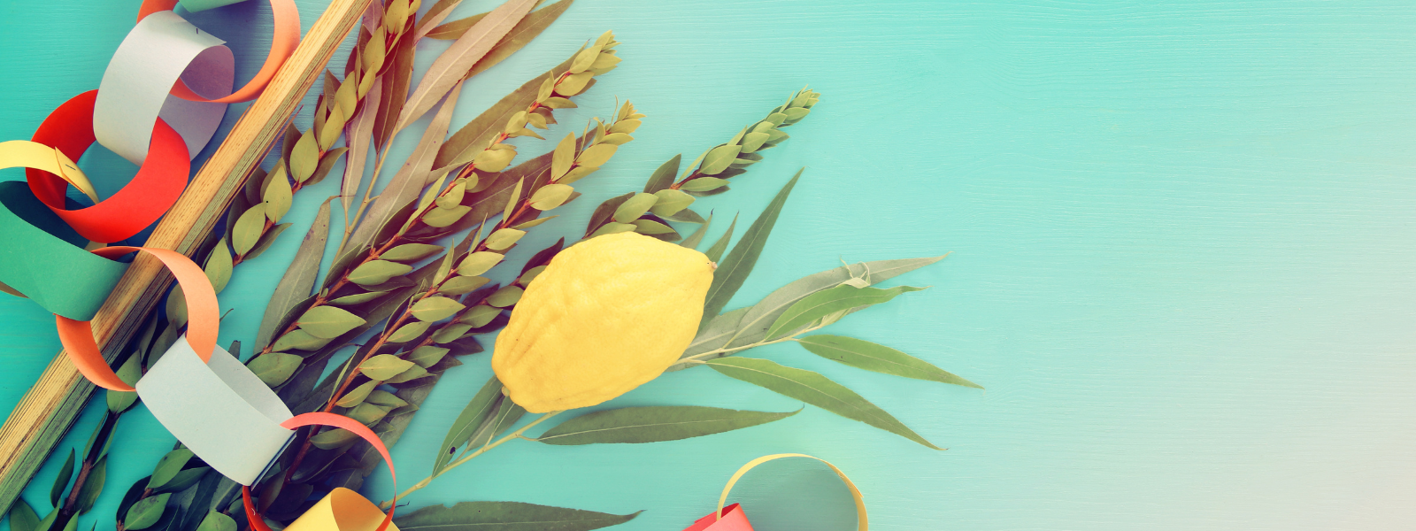 Colorful festive ribbons and a yellow citron are placed on green palm and myrtle branches against a light blue background, conveying a joyful mood.