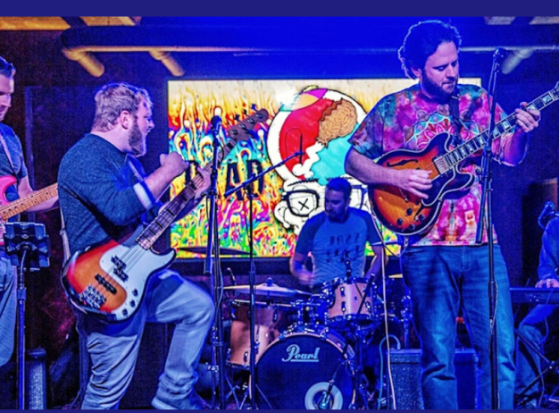 A five-member band performs on stage with vibrant, colorful lights in the background. The musicians play guitars, bass, drums, and keyboard, creating an energetic atmosphere.