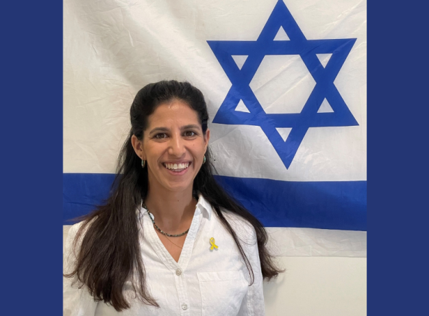 A woman in a white shirt, smiling, stands in front of an Israeli flag, featuring a blue Star of David. The mood is positive and proud.