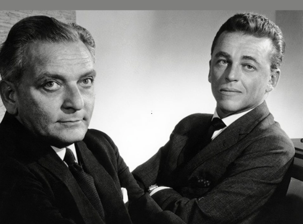 Two men in suits pose confidently in a black and white photo. One faces forward with a serious expression, while the other looks over, smiling slightly.