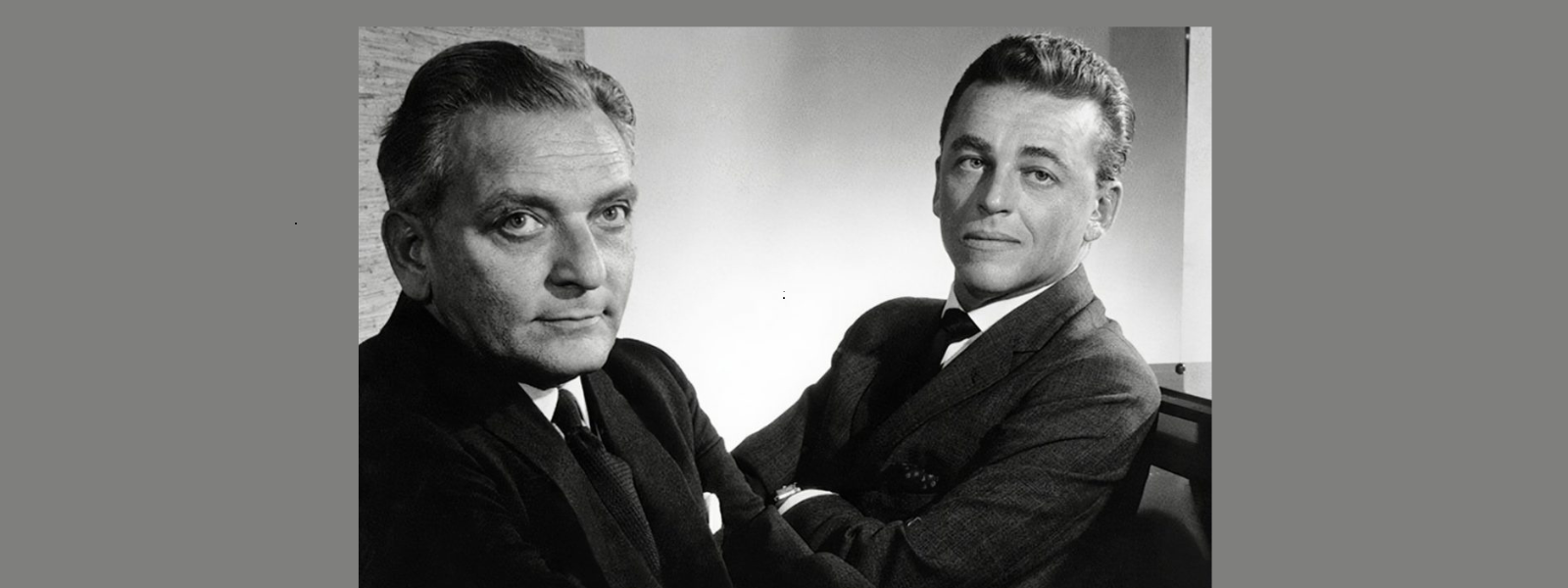 Two men in suits pose confidently in a black and white photo. One faces forward with a serious expression, while the other looks over, smiling slightly.