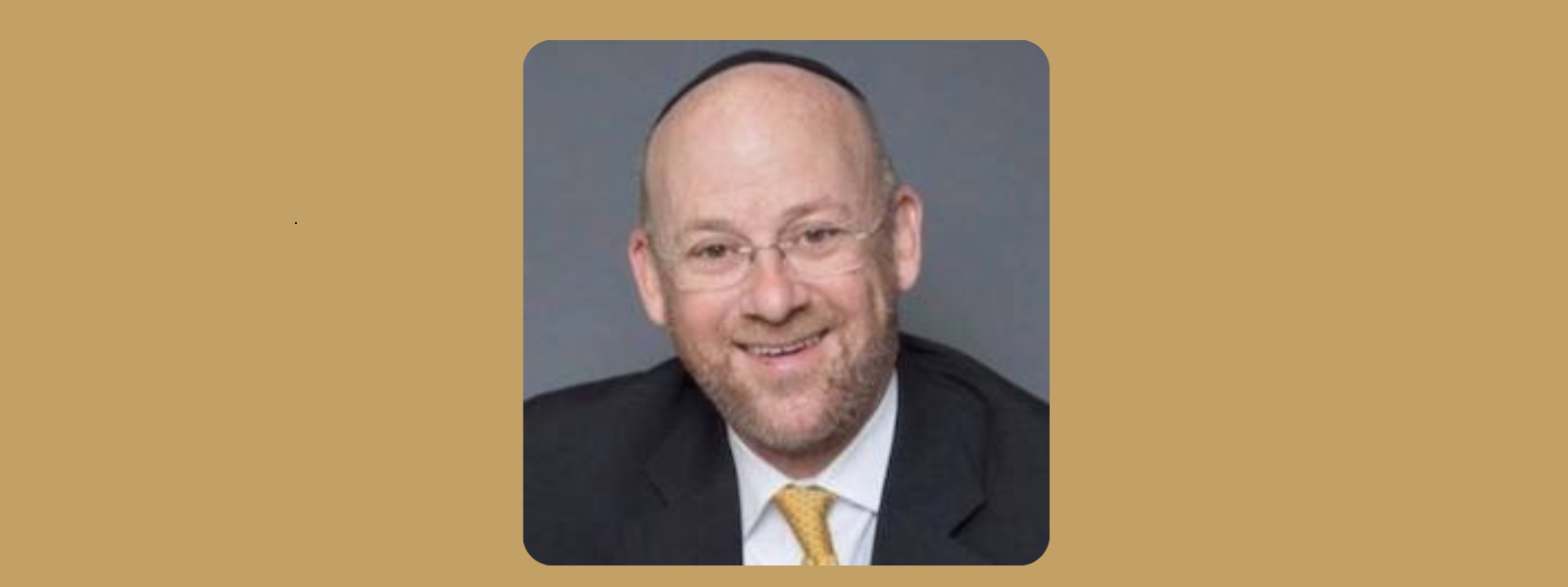 A smiling man with glasses, wearing a kippah and a suit with a yellow tie, set against a plain gray background. The mood is friendly and approachable.