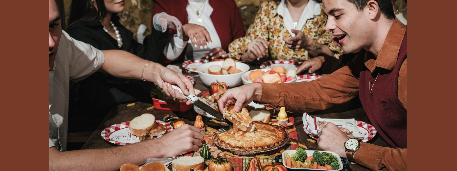 A group gathers around a festive table sharing pie, bread, and vegetables. Warm lighting and autumnal decor create a cozy, joyful atmosphere.