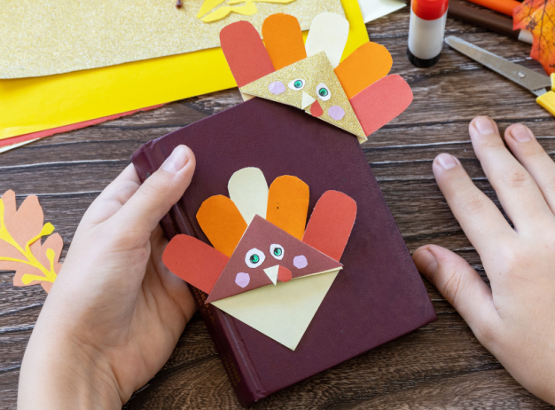 Two hands hold a notebook with a colorful turkey bookmark made from paper. Scissors, glue, and craft supplies are scattered on the wooden table.
