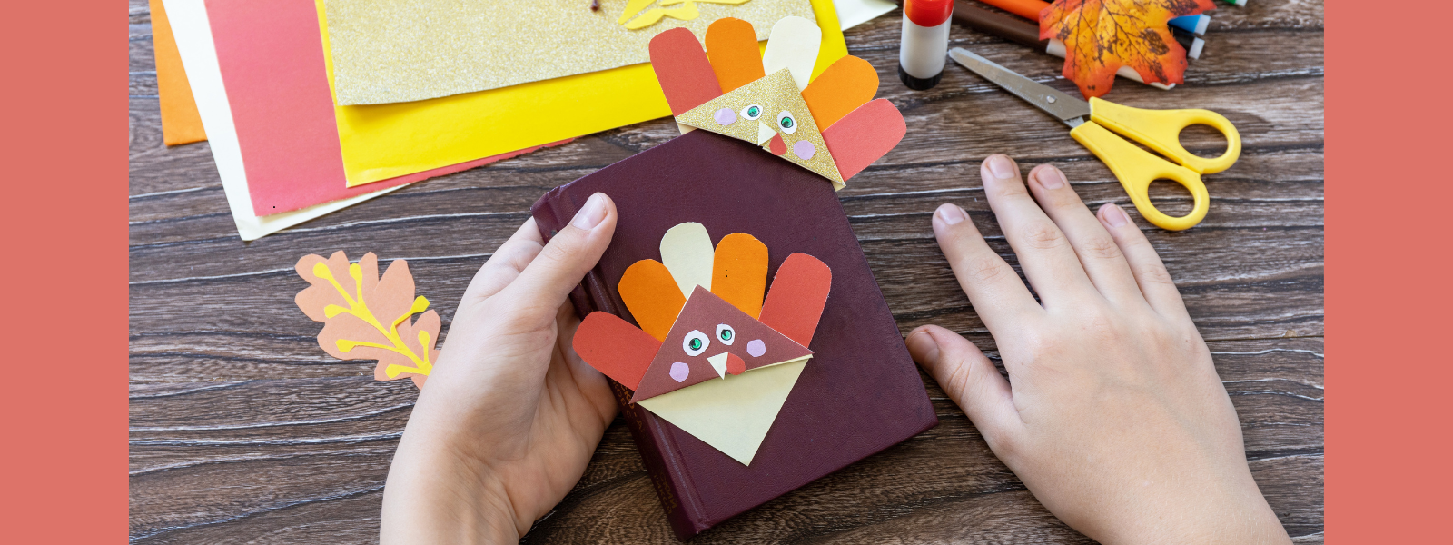 Two hands hold a notebook with a colorful turkey bookmark made from paper. Scissors, glue, and craft supplies are scattered on the wooden table.
