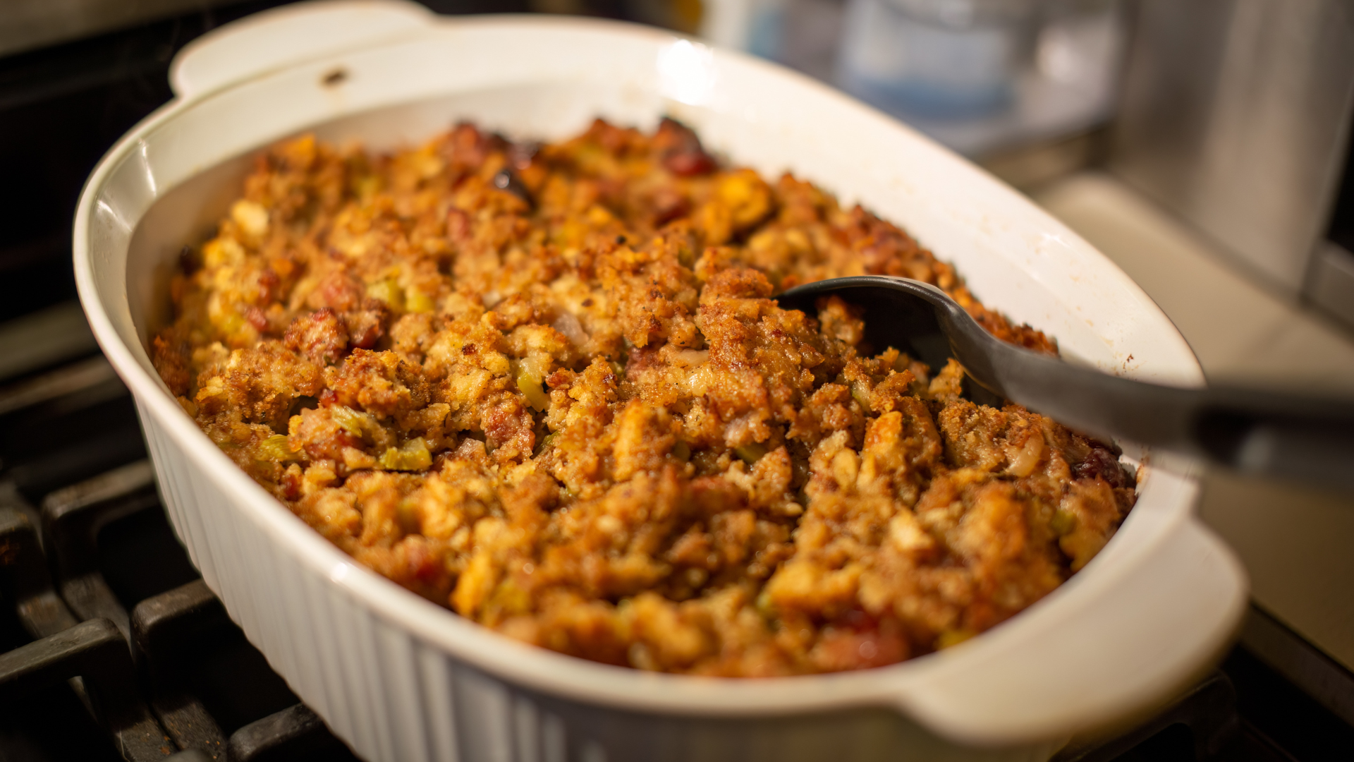 A white casserole dish filled with golden-brown stuffing on a stovetop. A serving spoon is poised to scoop, evoking a warm, home-cooked meal vibe.