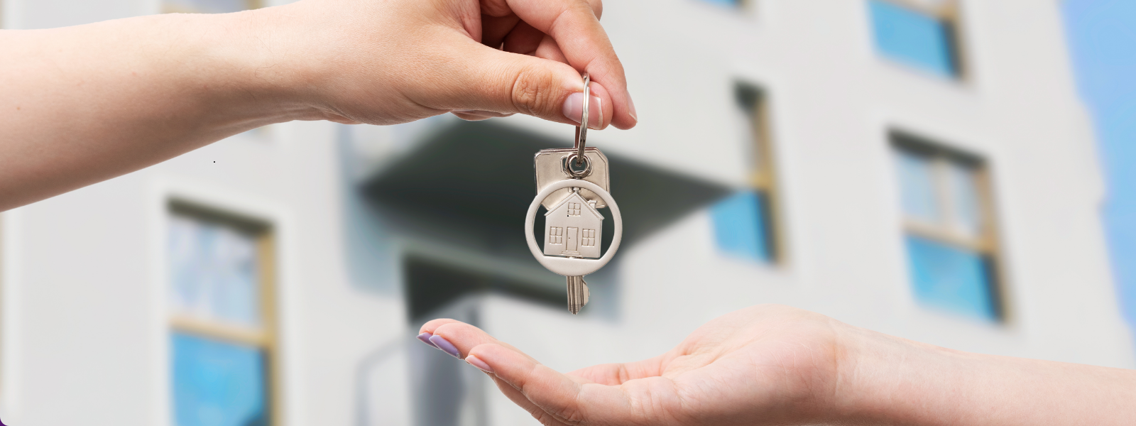 A hand holds out a key with a house-shaped keychain to another hand against a blurred building backdrop, conveying a sense of home ownership or rental.