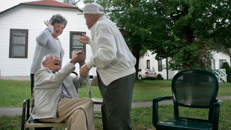 Three elderly people share a joyful moment outdoors. A woman smiles while two men laugh and hold hands, conveying happiness and warmth in a park-like setting.