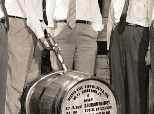 Five men in formal attire stand around a labeled bourbon barrel, observing it intently. The setting has a historical, industrial ambiance.