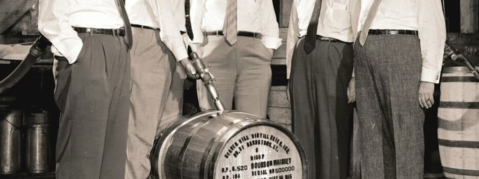 Five men in formal attire stand around a labeled bourbon barrel, observing it intently. The setting has a historical, industrial ambiance.