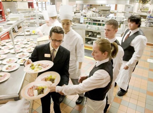 Restaurant kitchen scene with chefs and waitstaff in uniforms, preparing and serving dishes. The environment is busy yet orderly, conveying teamwork.