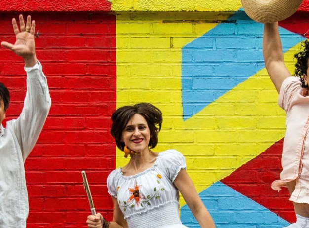 Three people in vibrant clothing jump joyfully in front of a colorful mural with red, yellow, and blue shapes. Two men hold hats, and a woman smiles. Energetic and lively.