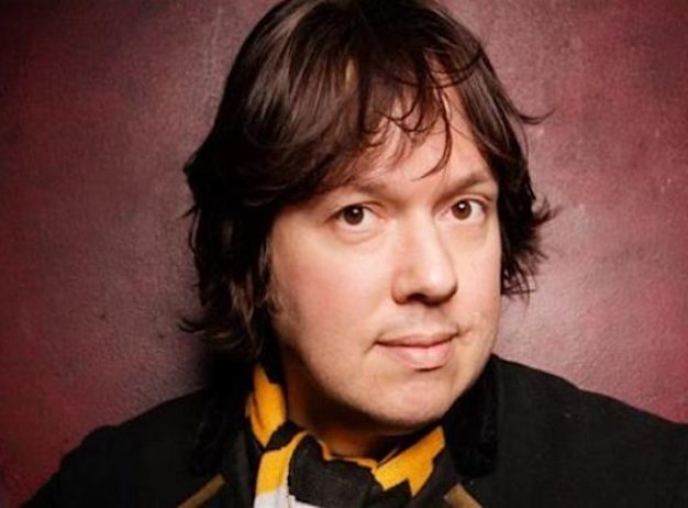 A person with medium-length dark hair looks directly at the camera, wearing a black coat and a yellow scarf. The background is a textured reddish wall, creating a warm tone.