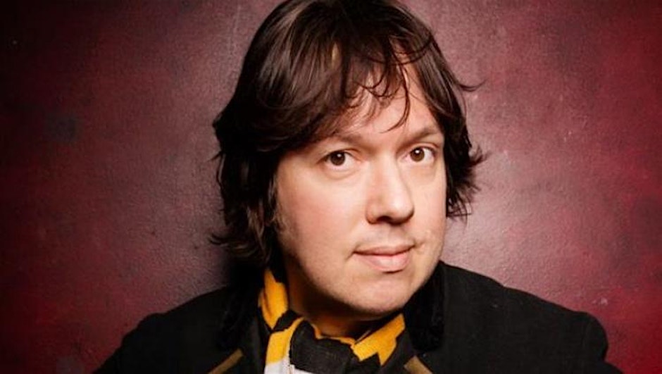 A person with medium-length dark hair looks directly at the camera, wearing a black coat and a yellow scarf. The background is a textured reddish wall, creating a warm tone.