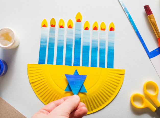 A hand crafts a Hanukkah menorah using a yellow paper plate and blue paper. Paintbrushes, scissors, and blue paint jars are on a light background.