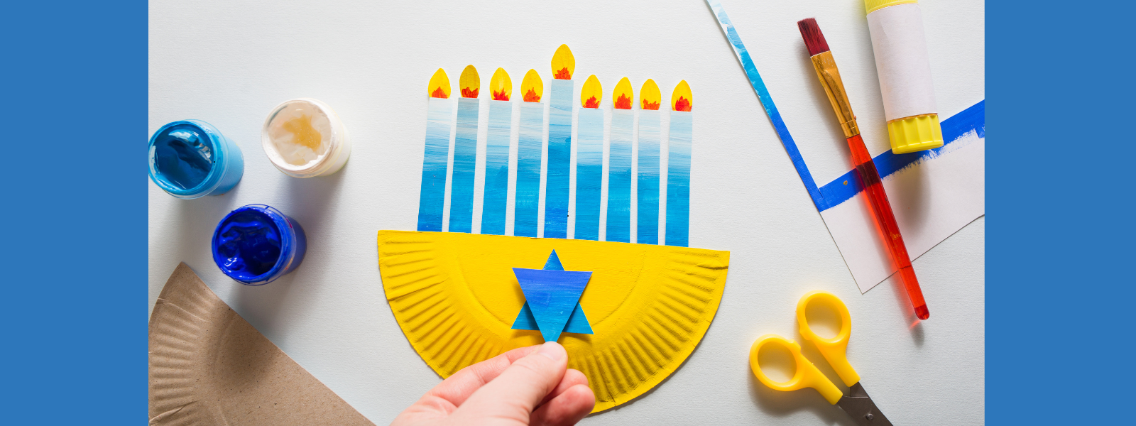 A hand crafts a Hanukkah menorah using a yellow paper plate and blue paper. Paintbrushes, scissors, and blue paint jars are on a light background.