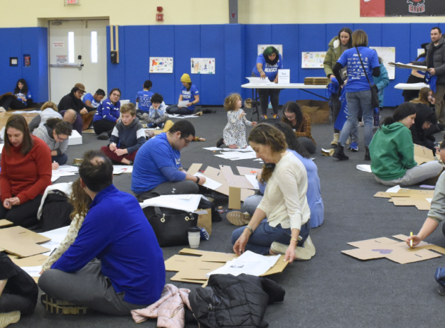 Group of families and children are crafting with cardboard on a gym floor. The atmosphere is lively, with colorful artwork on the walls and tables.