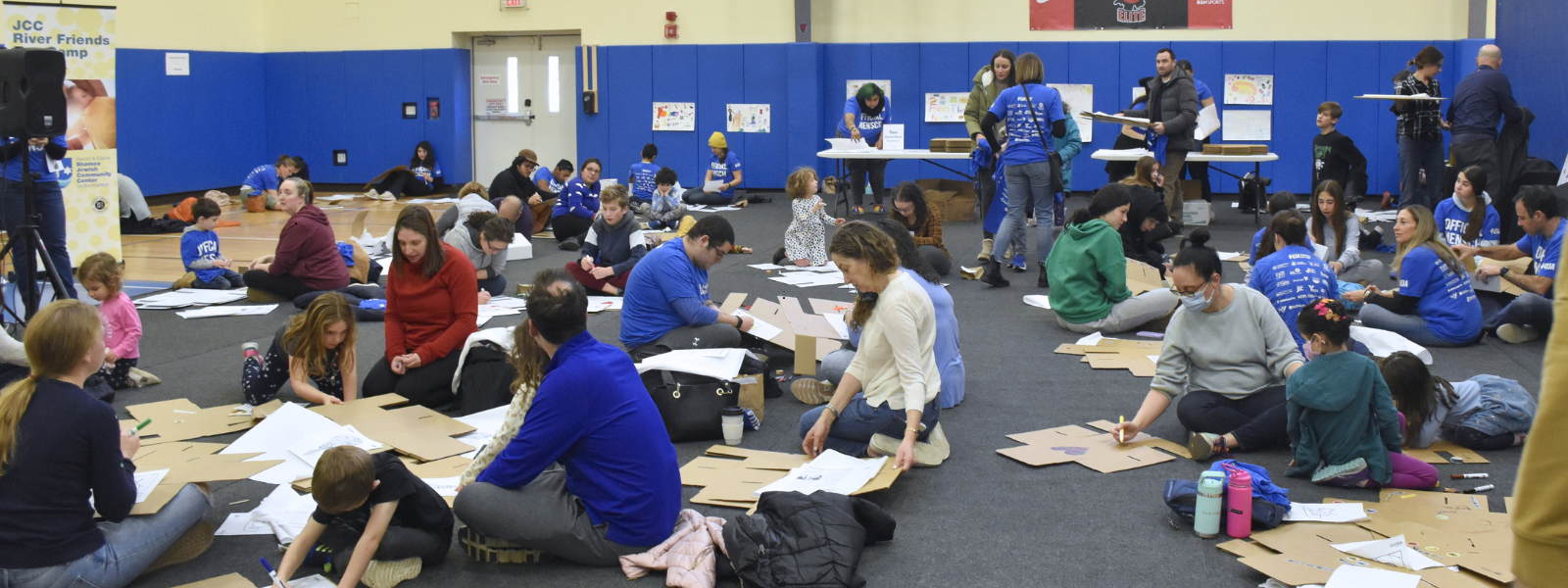 Group of families and children are crafting with cardboard on a gym floor. The atmosphere is lively, with colorful artwork on the walls and tables.