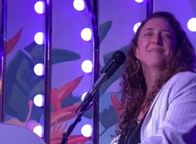 A person in a white jacket smiles while playing piano on a vibrant stage with purple lighting and round bulbs. The scene conveys joy and energy.