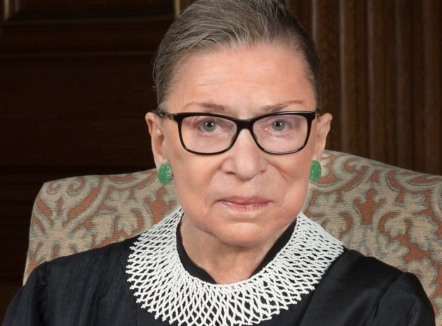 An older woman with glasses and a serious expression sits on a patterned chair, wearing a lace collar and green earrings, conveying strength and dignity.
