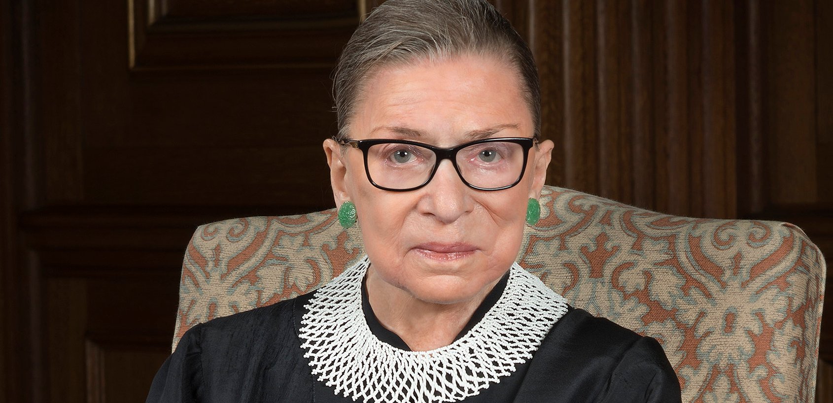 An older woman with glasses and a serious expression sits on a patterned chair, wearing a lace collar and green earrings, conveying strength and dignity.