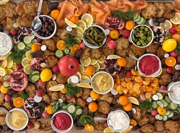 A vibrant spread of diverse foods on a table, including smoked salmon, sliced cucumbers, various dips, citrus slices, and small fruits. The tone is festive and colorful.