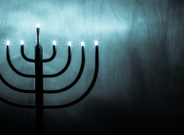 A menorah with seven lit candles stands against a dark, textured background. The soft glow creates an atmosphere of serenity and reflection.