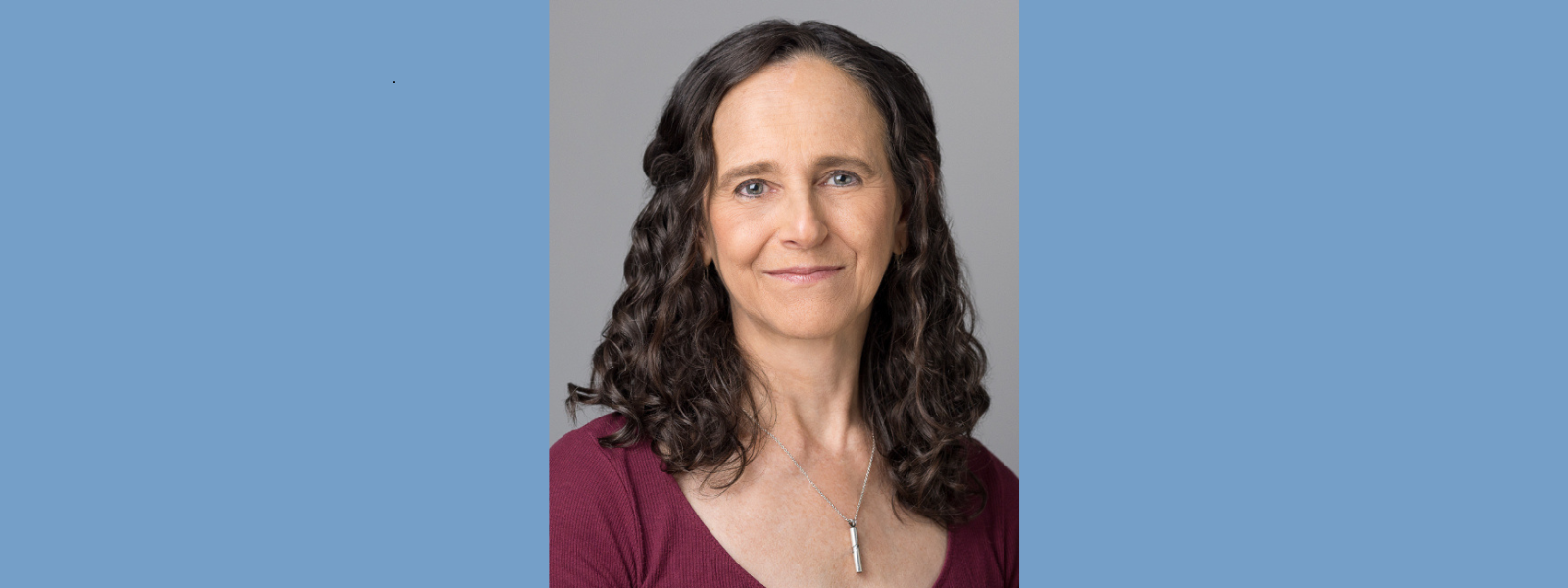 A woman with curly dark hair smiles softly against a neutral gray background. She wears a maroon top and a pendant necklace, conveying a calm, approachable presence.