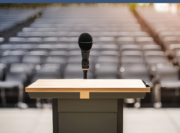 A microphone stands on a podium in an empty auditorium. Rows of blurred gray seats are in the background, conveying a sense of anticipation.