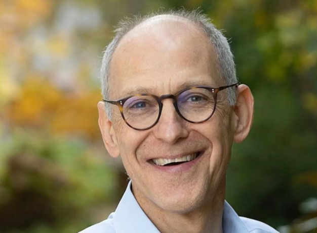 A smiling older man wearing glasses and a light blue shirt is outdoors, with a blurred background of autumnal trees. The mood is warm and approachable.