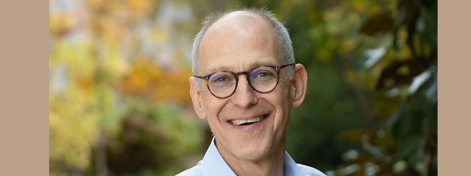 A smiling older man wearing glasses and a light blue shirt is outdoors, with a blurred background of autumnal trees. The mood is warm and approachable.