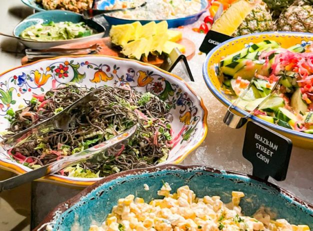 A vibrant buffet table features colorful dishes like Brazilian street corn, salad, and fruit garnished with pineapples. Chefs in white uniforms serve guests.