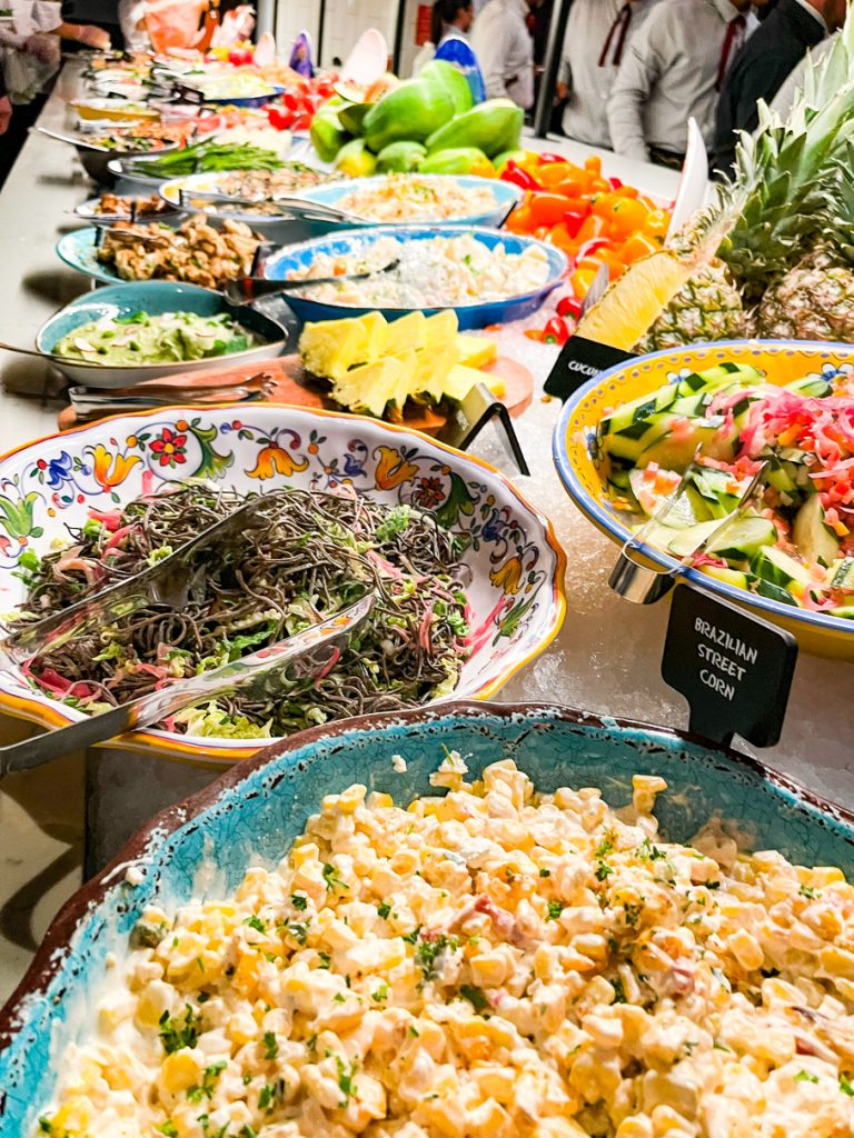 A vibrant buffet table features colorful dishes like Brazilian street corn, salad, and fruit garnished with pineapples. Chefs in white uniforms serve guests.