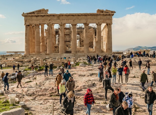 A large crowd of tourists explores the ancient ruins of the Parthenon under a partly cloudy sky. The scene is lively, with people walking and taking photos.