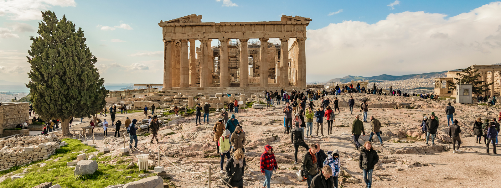 A large crowd of tourists explores the ancient ruins of the Parthenon under a partly cloudy sky. The scene is lively, with people walking and taking photos.