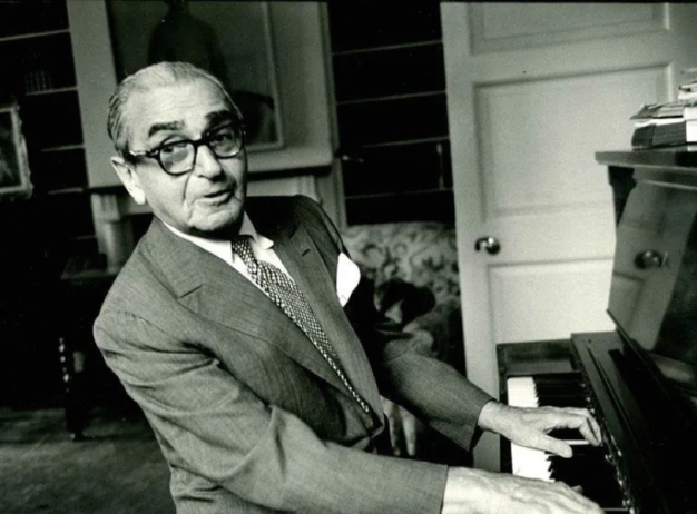 Black and white photo of an older man playing the piano in a cozy room. He wears glasses and a suit, exuding a joyful and nostalgic mood.