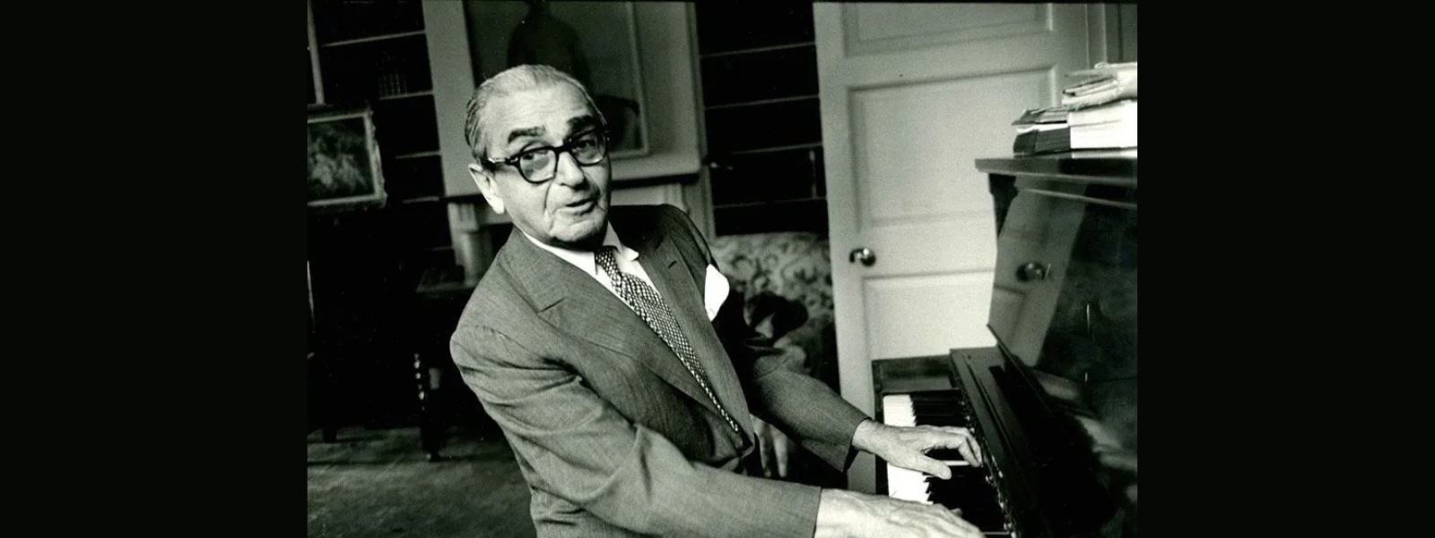 Black and white photo of an older man playing the piano in a cozy room. He wears glasses and a suit, exuding a joyful and nostalgic mood.