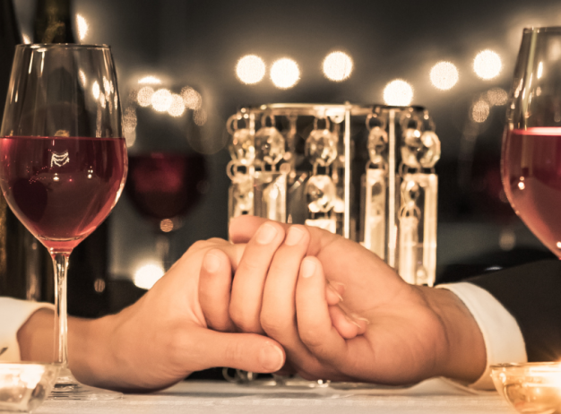 A romantic dinner setting with two hands clasped over a white tablecloth, flanked by glasses of red wine. Warm candlelight creates a cozy, intimate ambiance.