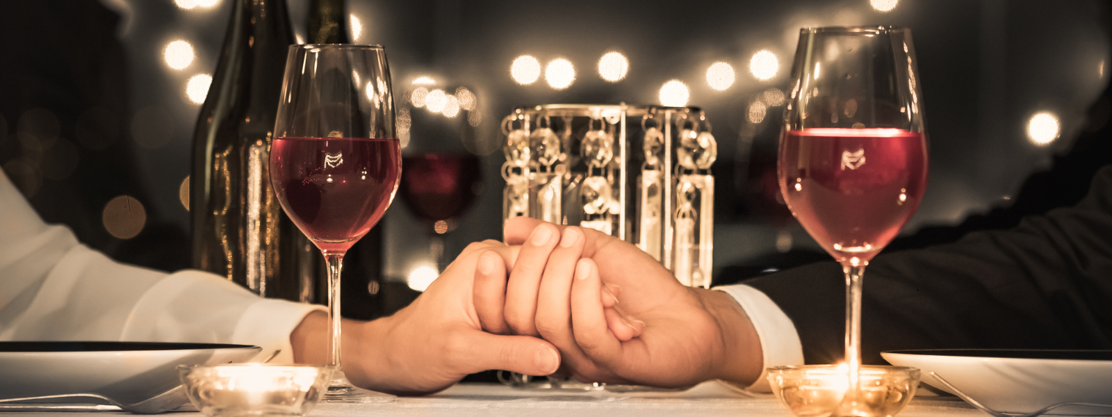 A romantic dinner setting with two hands clasped over a white tablecloth, flanked by glasses of red wine. Warm candlelight creates a cozy, intimate ambiance.