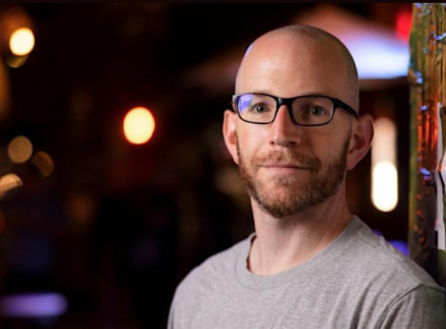 Bald man with glasses and a beard, wearing a gray T-shirt, stands against a vibrant graffiti wall at night. Dim lights create a warm, relaxed ambiance.