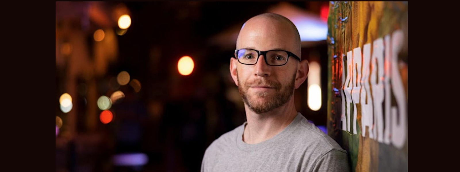 Bald man with glasses and a beard, wearing a gray T-shirt, stands against a vibrant graffiti wall at night. Dim lights create a warm, relaxed ambiance.