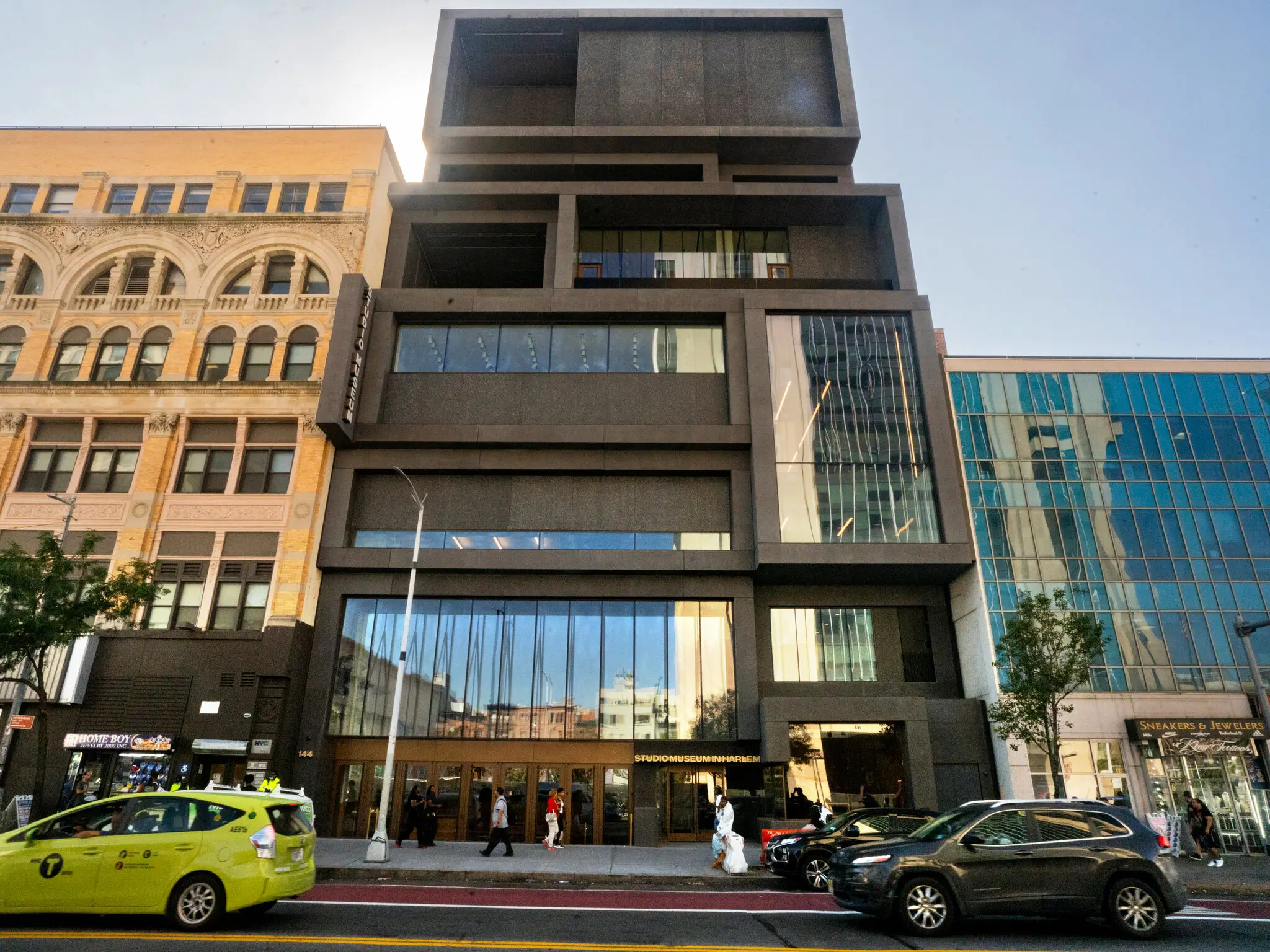 A modern, geometric black building with large glass windows stands between a classic beige building and a reflective glass structure. Taxis and pedestrians pass by.