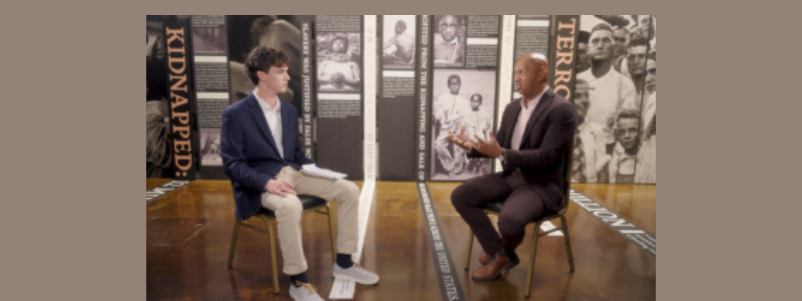 Two people sit on chairs engaged in conversation in a museum setting. Behind them are panels with historical images and text. The atmosphere is serious.